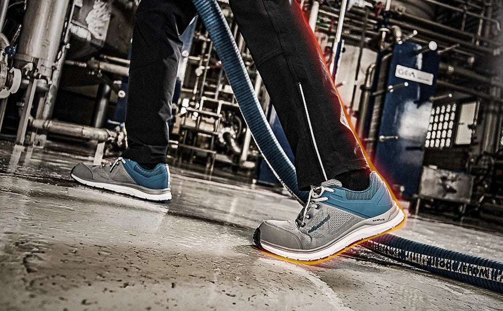 Sicherheitsschuh ‚Lift Impulse‘ von Albatros in einer industriellen Umgebung, getragen von einer Person auf nassem Boden mit reflektierender Markierung und blauen Schläuchen im Hintergrund.