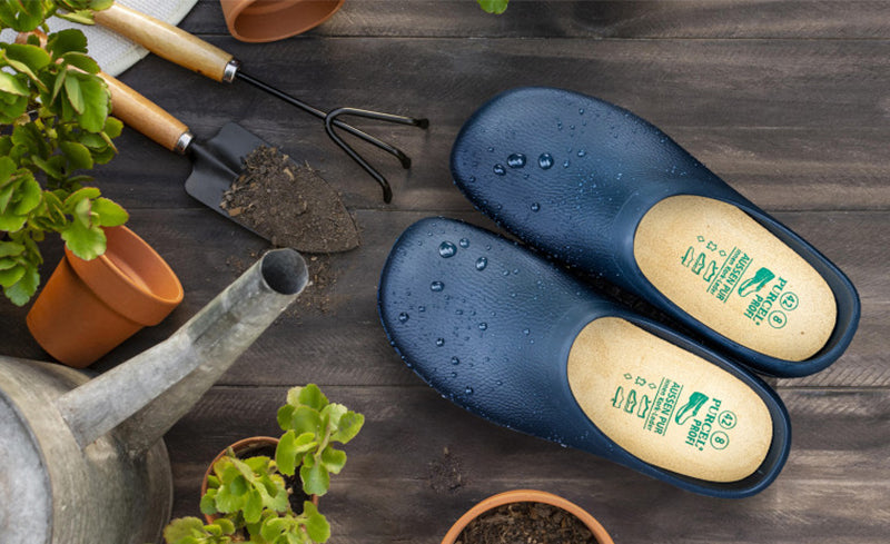 Ein Paar navyblaue "Purcel" Gartenclogs mit Wassertropfen auf der Oberfläche, arrangiert auf Holztisch mit Gartengeräten, Gießkanne, Blumentöpfen und frischem Grün – Szenerie für Gartenarbeit.