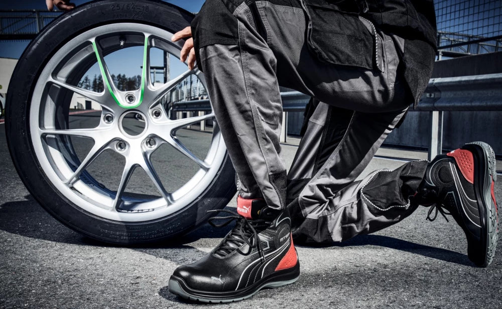 Sicherheitsschuh "Touring" von Puma Safety - Person kniet auf Asphalt neben einer Rennfelge, moderner Arbeitsschuh mit schwarzem Obermaterial und roten Akzenten.