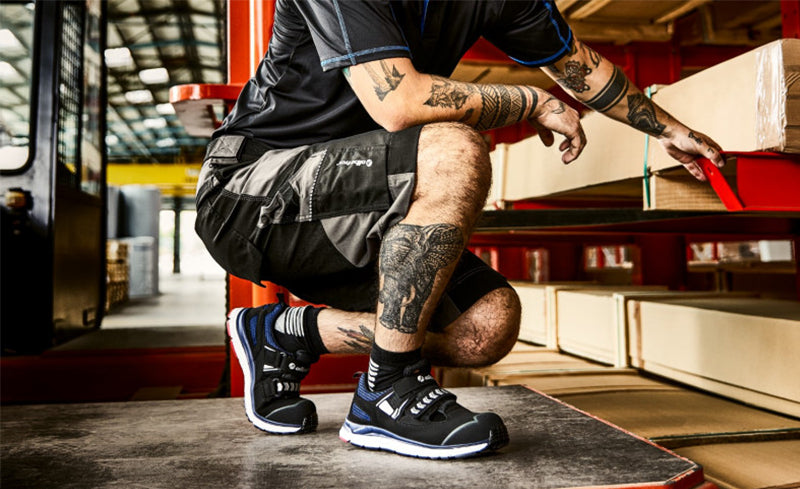 Ein tätowierter Lagerarbeiter kniet beim Kommissionieren in einem Hochregallager und trägt die schwarze Sicherheitssandale ‚Jetstream‘ von Albatros mit weißer Sohle.