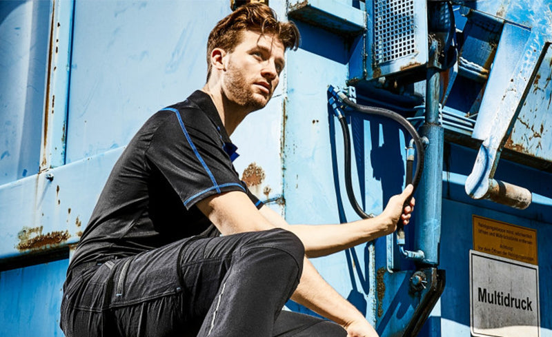 Mann in schwarzer Arbeitskleidung mit blauen Akzenten sitzt vor einer blauen Industrieanlage und hält sich an einer Metallstange fest, im Hintergrund ein Schild mit der Aufschrift "Multidruck".