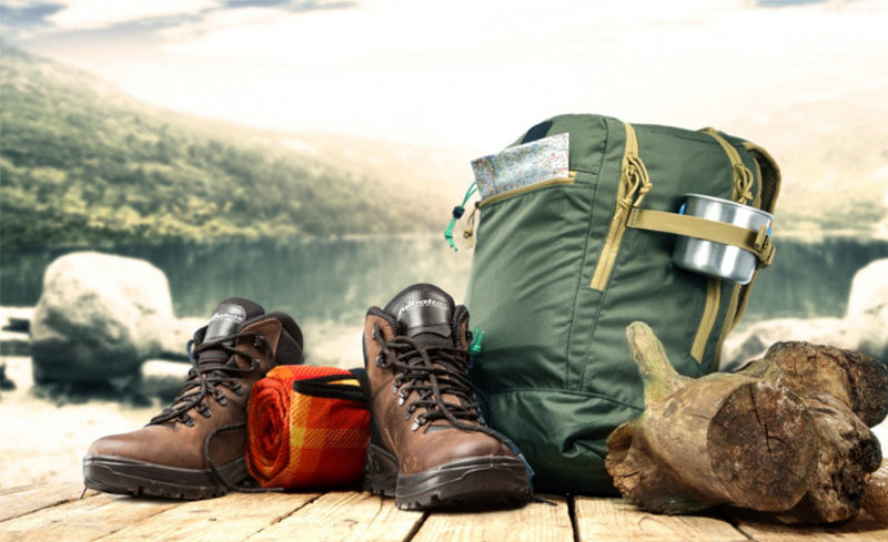 Wanderausrüstung mit Wanderschuhen, Rucksack, Decke und Trinkbecher auf Holzplattform vor Bergsee.
