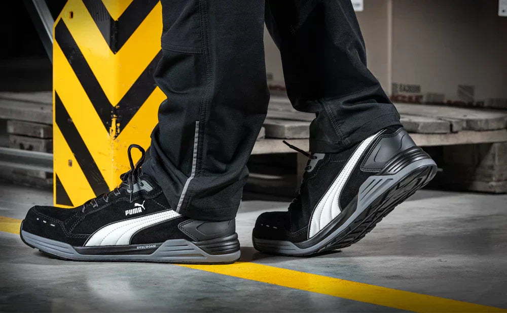 Sicherheitsschuh "Airtwist" von Puma Safety in Schwarz mit weißem Puma-Logo, getragen von einer Person in schwarzer Arbeitskleidung auf einem Industrieboden mit gelber Markierung.