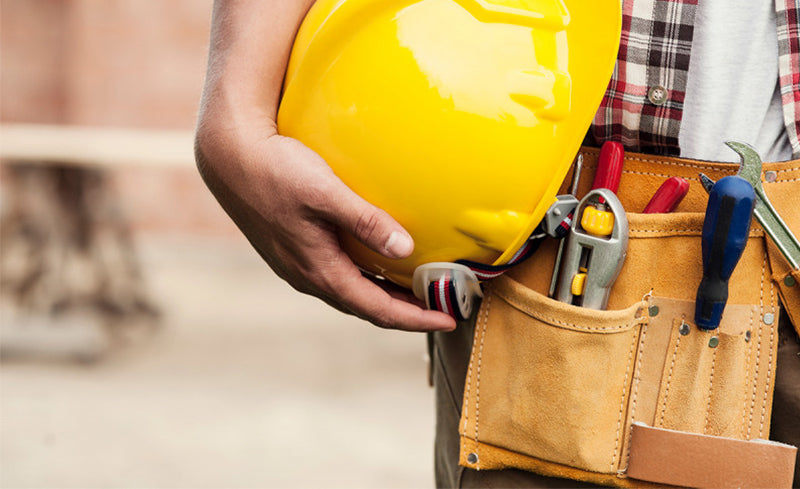 Nahaufnahme eines Bauarbeiters mit gelbem Schutzhelm in der Hand und Werkzeuggürtel voller Werkzeuge um die Hüfte.