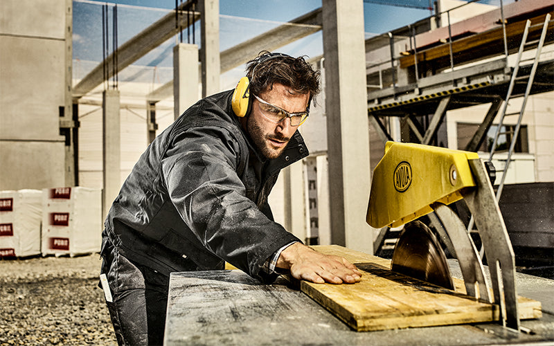 Handwerker mit Gehörschutz und Schutzbrille schiebt Holz durch eine Kreissäge auf einer Baustelle.