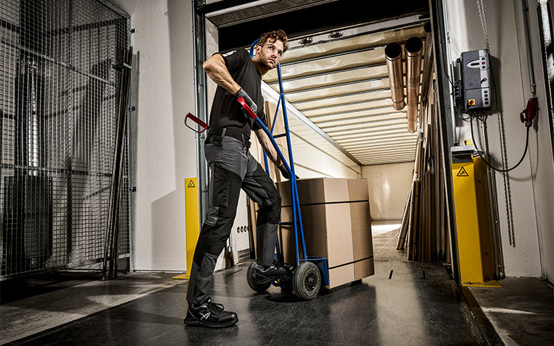 Lagermitarbeiter trägt Arbeitskleidung und schiebt eine Sackkarre mit Kartons beim Beladen eines LKWs im Lager.