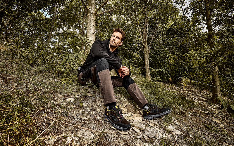 Mann in brauner Outdoorkleidung sitzt im Wald auf einem Hang und trägt robuste Wanderschuhe von Albatros.