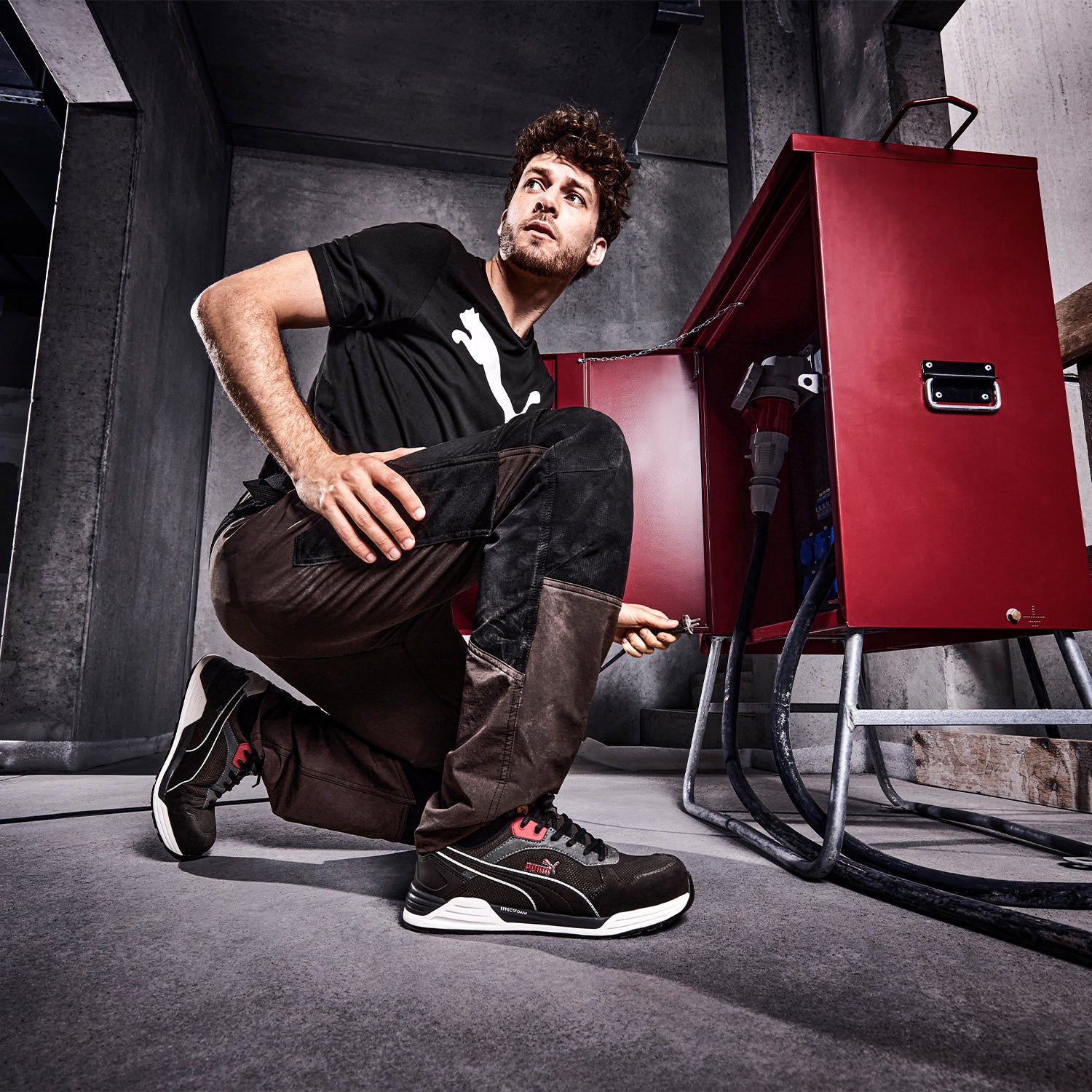 Mann in einem schwarzen T-Shirt von Puma und brauner Arbeitshose von Albatros kniet vor einem offenen roten Werkzeugschrank, hält einen Kabel in der Hand und trägt die "Frontside Ivy" Sicherheitsschuhe von Puma Safety.