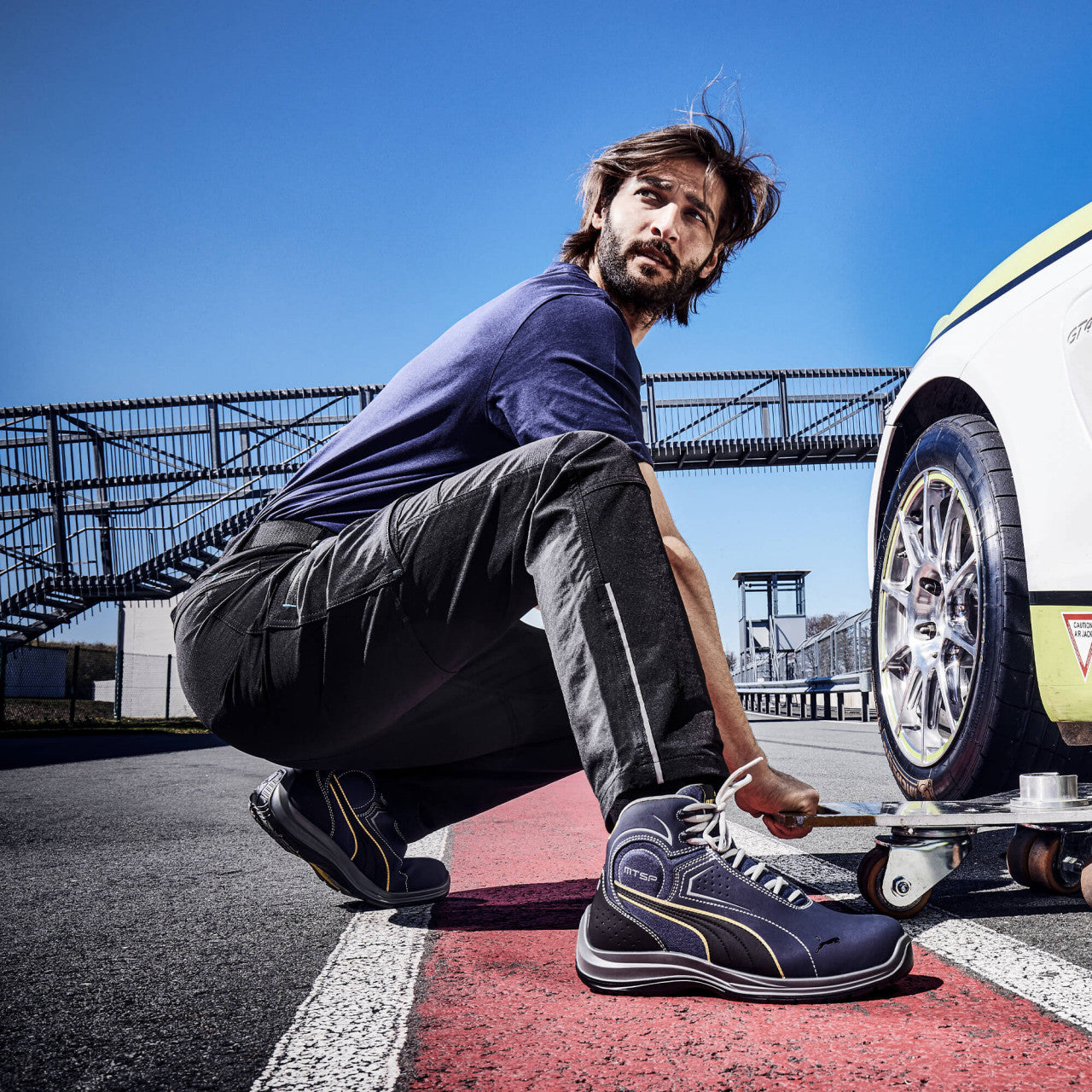 Ein Mann arbeitet auf einer Rennstrecke an einem Fahrzeug und trägt den Sicherheitsschuh "Touring" von Puma Safety in Blau mit gelben Details.