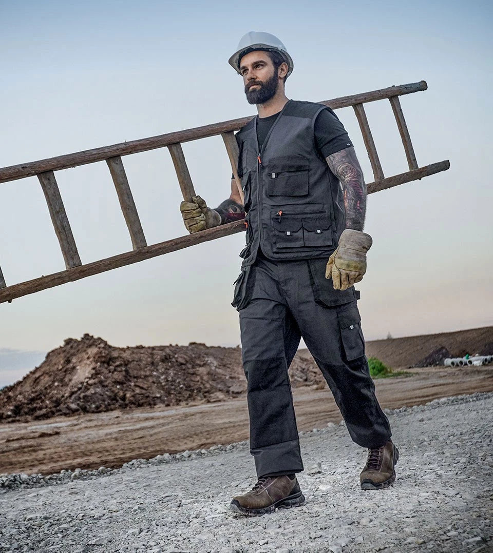 Bauarbeiter mit Schutzhelm und Arbeitshandschuhen trägt eine Holzleiter über der Schulter – bekleidet mit schwarzer Arbeitskleidung und den braunen Sicherheitsschuhen "Condor" von Puma Safety, im Hintergrund eine Baustelle mit Erdhügeln.