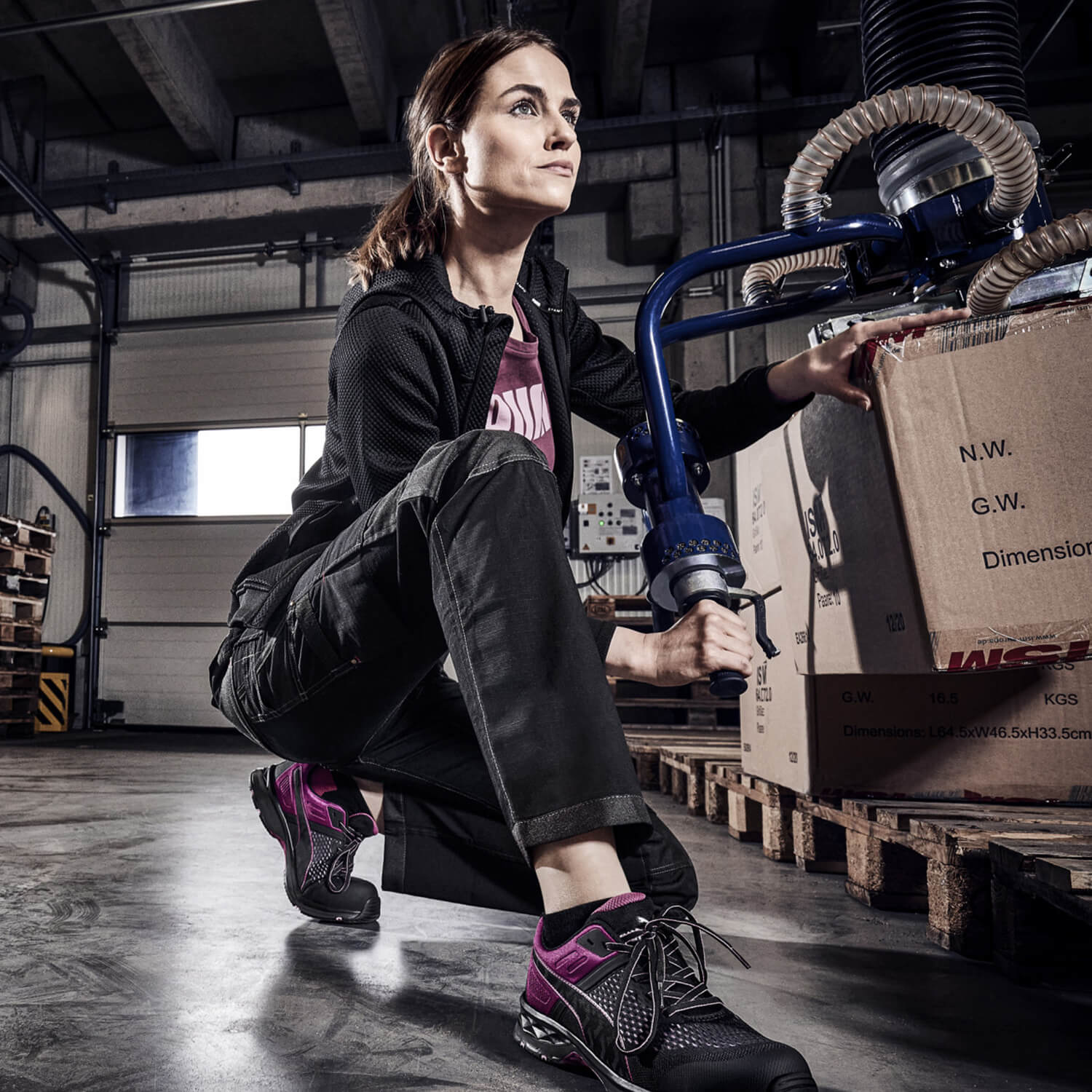 Eine Frau arbeitet in einer Lagerhalle mit einem Vakuumhebegerät und trägt den Sicherheitsschuh "Define" von Puma Safety in Schwarz und Magenta.