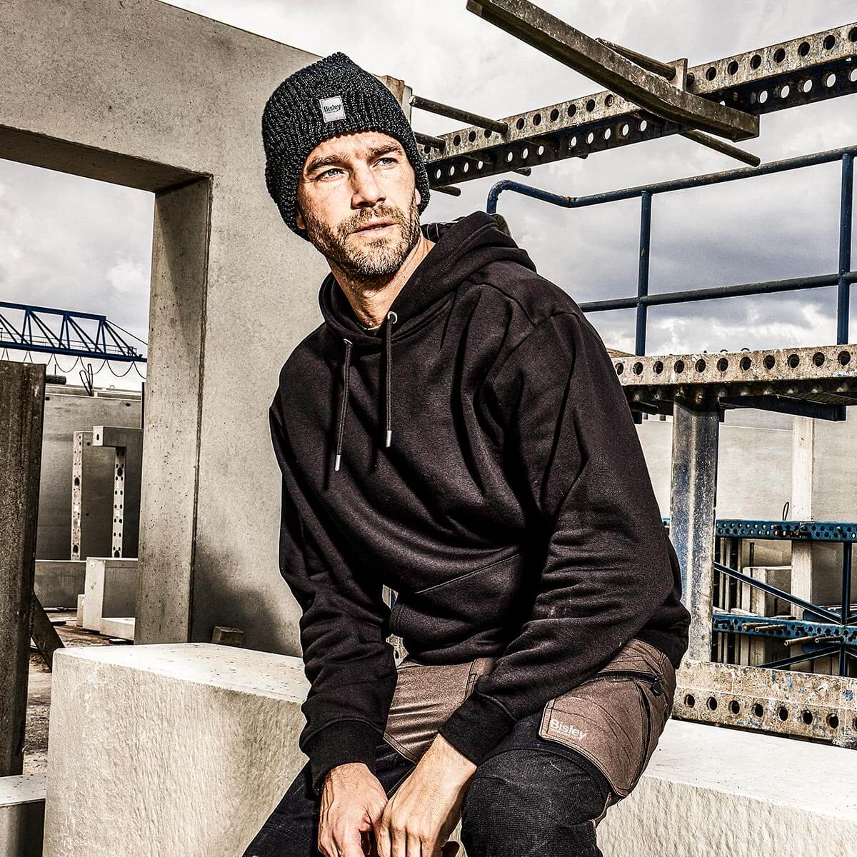 Sitzende Person mit Kapuzenpullover und Mütze von Bisley auf einer Baustelle mit Beton- und Metallkonstruktionen