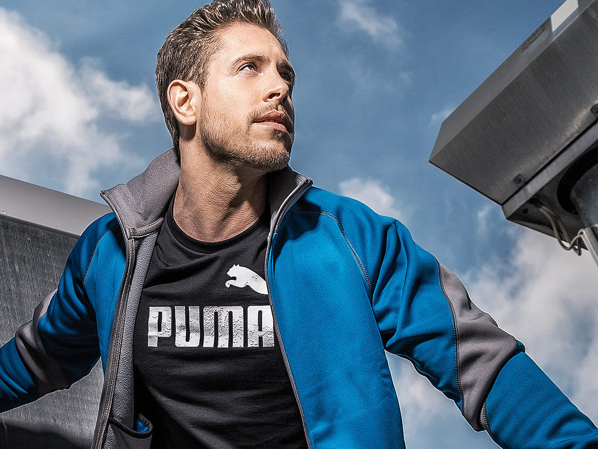 Mann mit blau-grauer Arbeitsjacke und schwarzem Puma T-Shirt blickt entschlossen nach oben, Hintergrund mit Himmel und technischer Anlage.