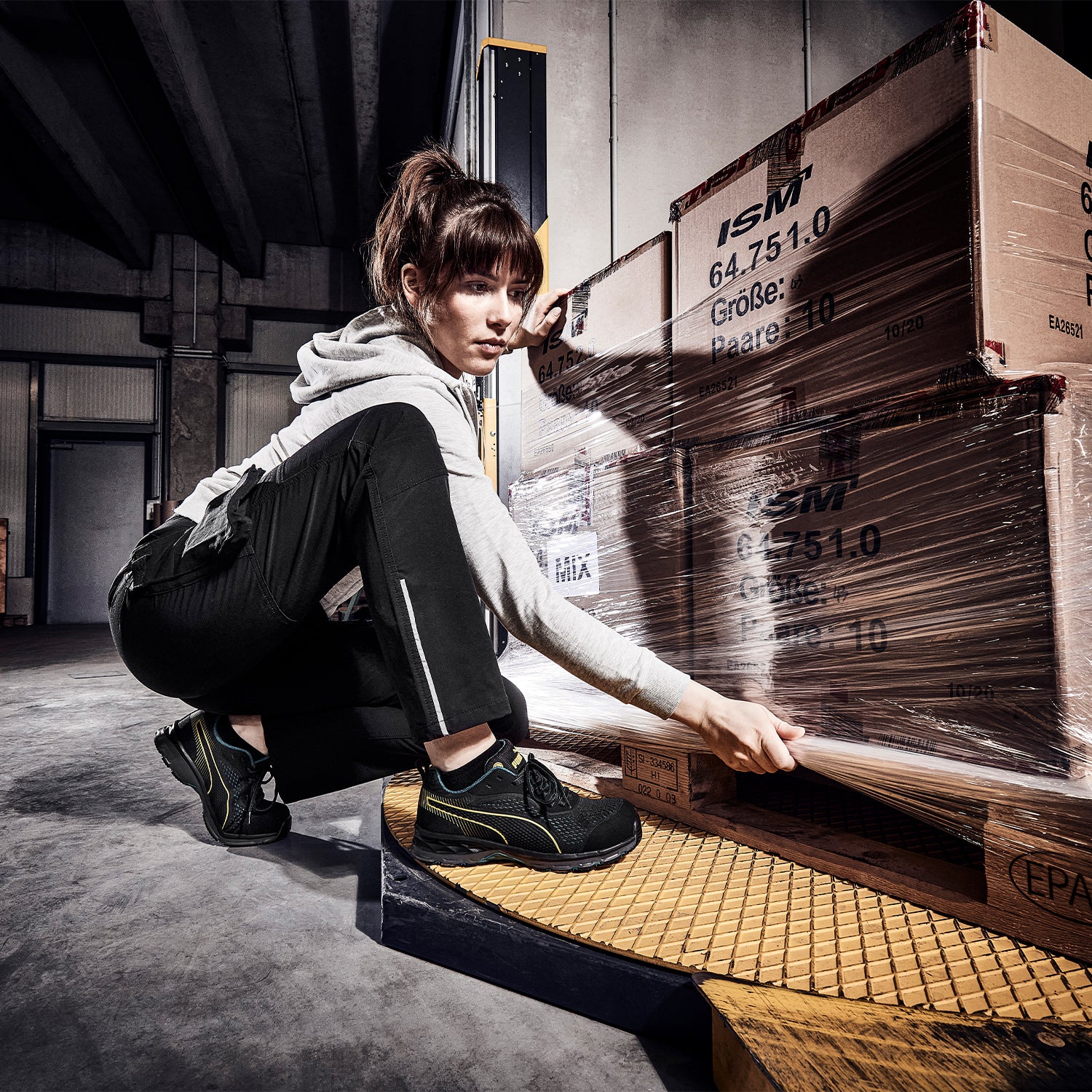 PUMA SAFETY FUSE KNIT WNS LOW Sicherheitsschuh S1P ESD in Schwarz mit gelben Akzenten, getragen von einer Person in hockender Position beim Verpacken einer Palette mit Stretchfolie in industrieller Lagerumgebung