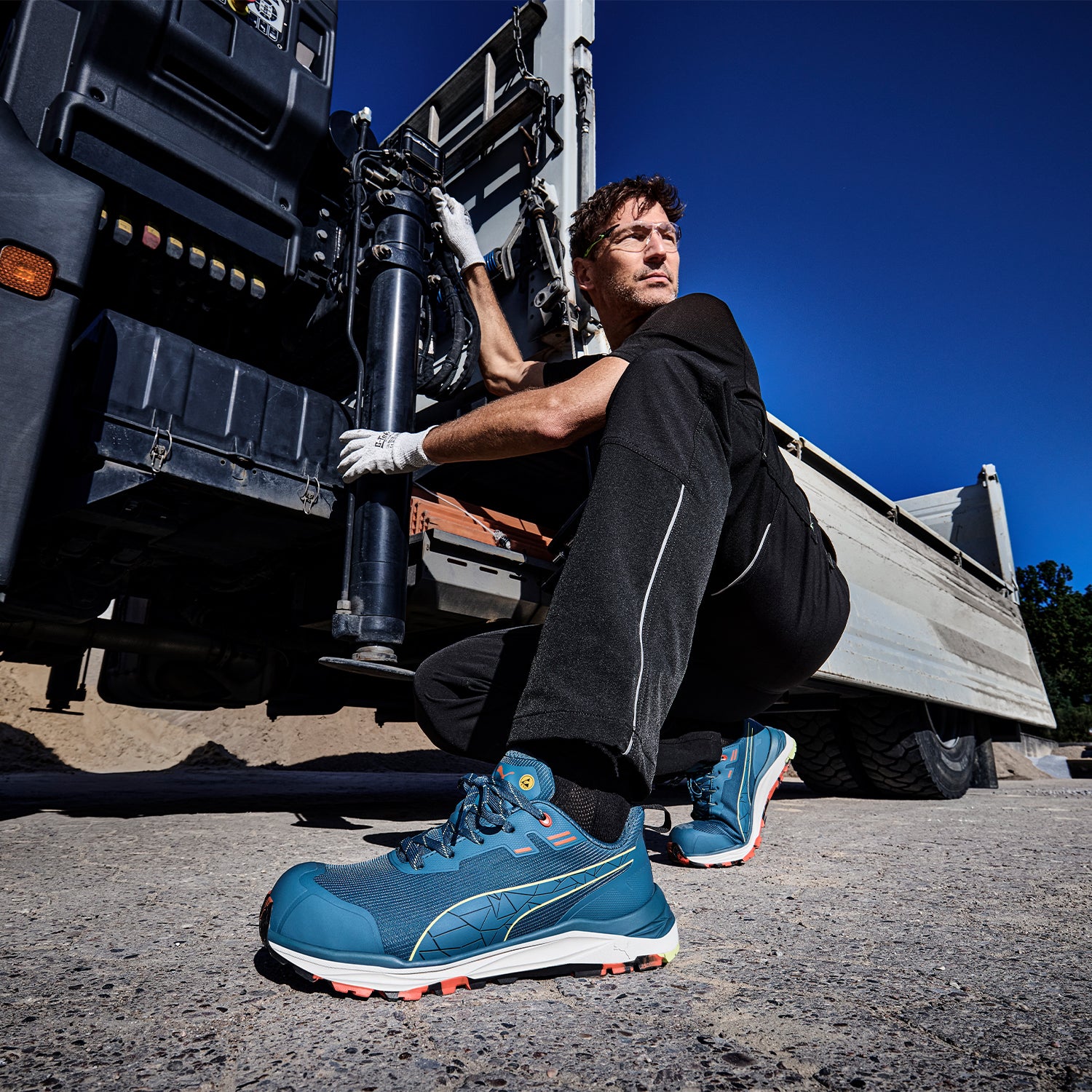 PUMA SAFETY XPLORE LOW Sicherheitsschuh S3S ESD in Blau mit grauen und orangefarbenen Akzenten, getragen von einer Person in kniender Position neben einem LKW bei Wartungsarbeiten im Außenbereich auf Asphaltboden