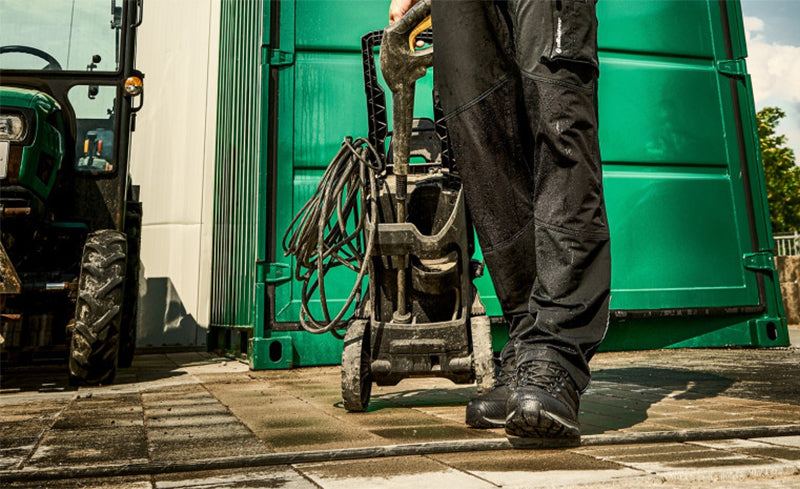Person in schwarzer Arbeitskleidung zieht einen Hochdruckreiniger über gepflasterten Boden, im Hintergrund grünes Tor und Traktor.