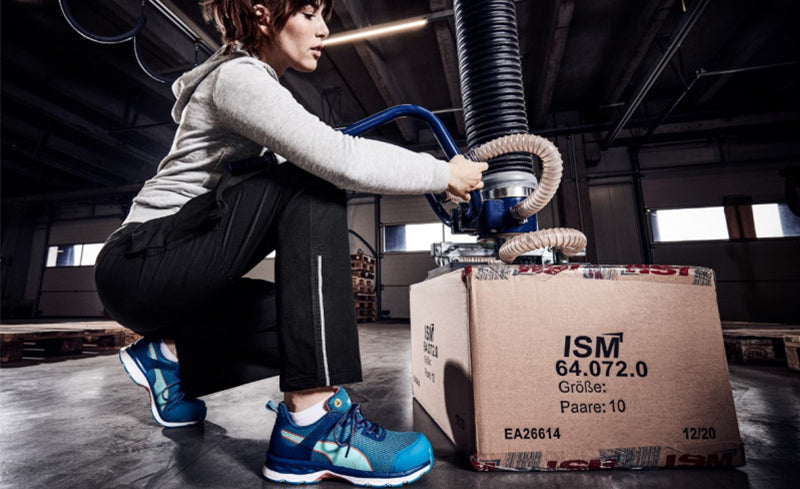 Frau mit dem blauen Sicherheitsschuh "Beat Wns" von PUMA Safety bei der Arbeit in einer Lagerhalle – hebt mithilfe eines Hebesystems einen Karton mit ISM-Beschriftung.