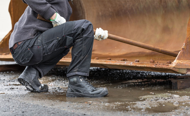 Arbeiter kniet auf nassem Boden vor einer rostigen Baggerschaufel und trägt den wetterfesten, schwarzen Sicherheitsschuh "Conquest" von Puma Safety.
