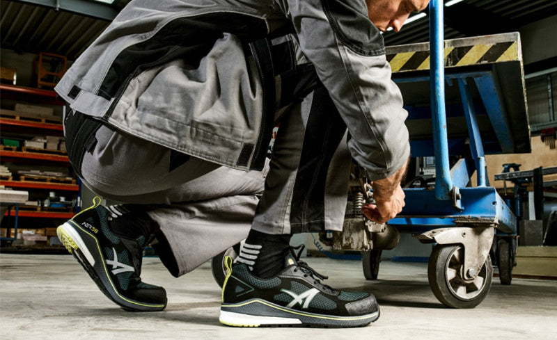 Arbeiter in grauer Arbeitskleidung kniet am Rollwagen in Werkhalle, trägt den Sicherheitsschuh "AER58" von Albatros.