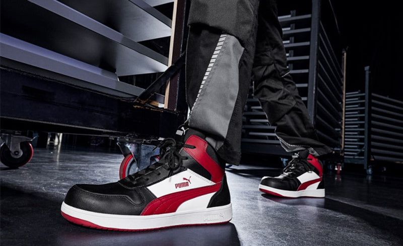 Arbeiter in schwarzer Arbeitshose schiebt Rollregal in Werkstatt, trägt den Sicherheitsschuh "Frontcourt" von Puma Safety in Schwarz-Rot-Weiß.