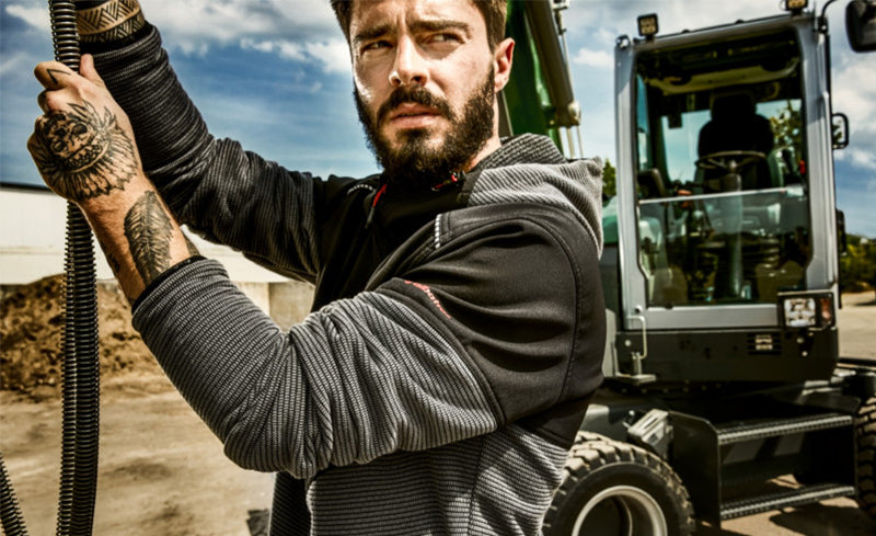 Mann mit Bart und Tätowierungen in Arbeitskleidung von Albatros hält ein Kabel vor einer Baumaschine auf einer Baustelle.
