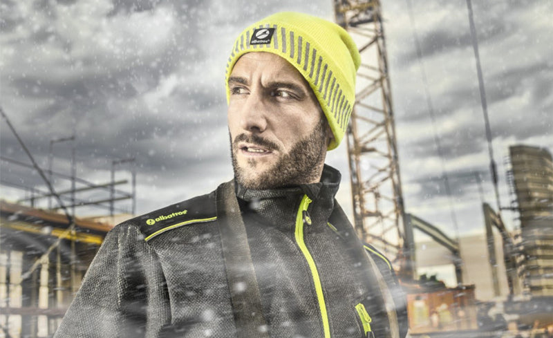 Bauarbeiter mit gelb-neonfarbener Mütze von Albatros und wetterfester Arbeitskleidung steht bei Schneetreiben vor einer Baustelle mit Kran.