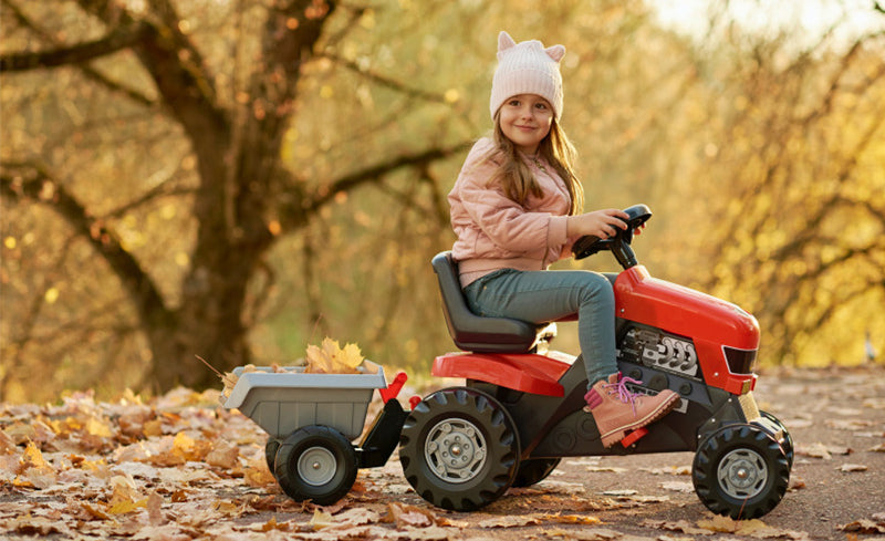Lächelndes Mädchen mit Mütze fährt im Herbst auf einem roten Trettraktor mit Anhänger voller Blätter durch einen Park.