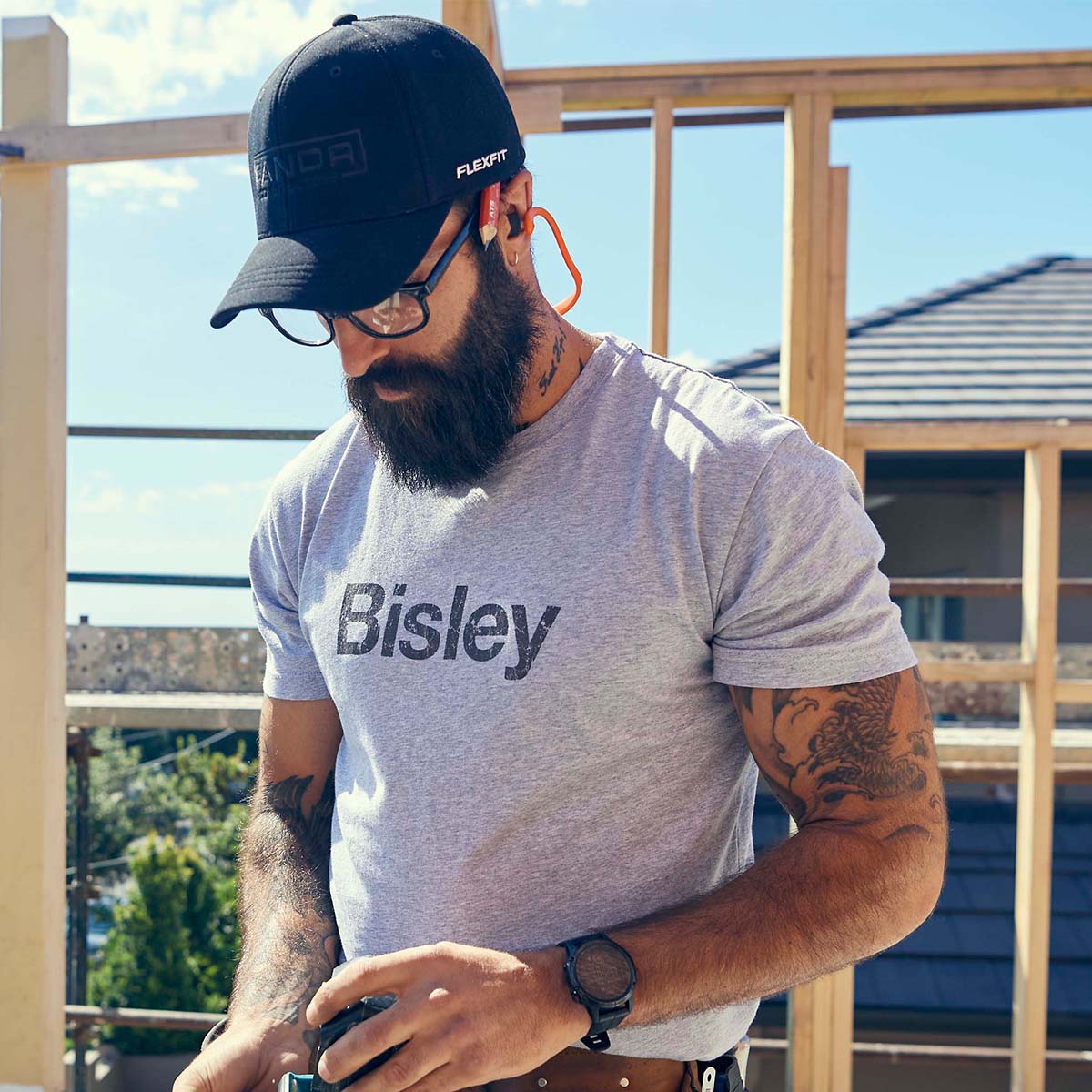 Handwerker mit Bart trägt Bisley Arbeitskleidung – graues T-Shirt, Schutzbrille und Cap – beim Arbeiten auf der Baustelle. Hochwertige, robuste Workwear für Profis.