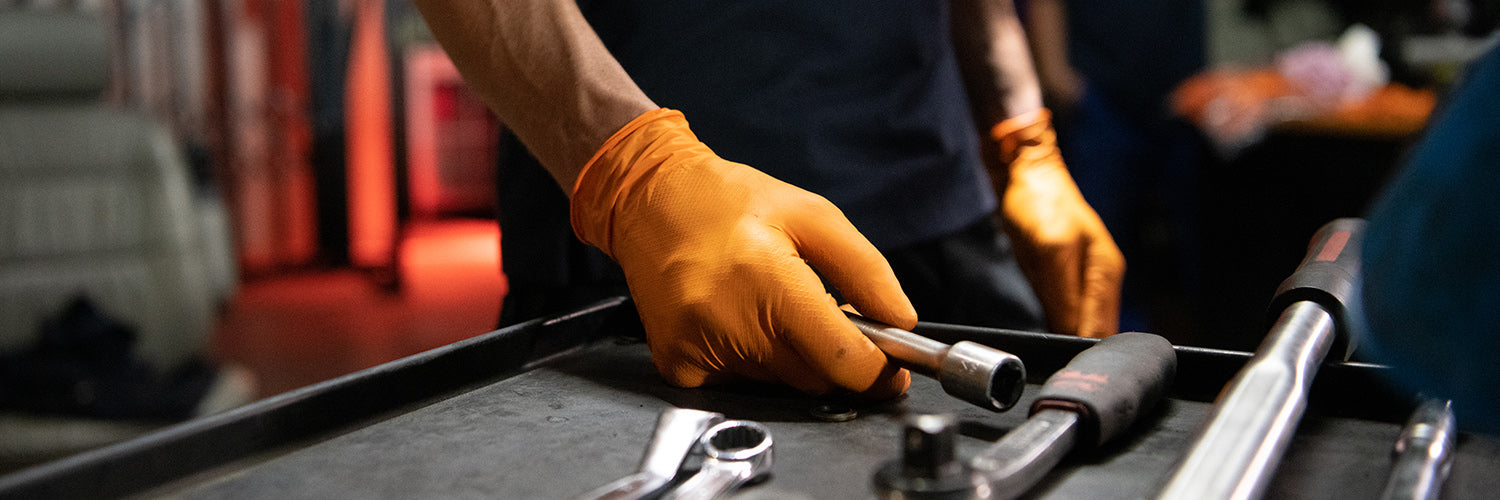 Person mit orangefarbenen PIP-Schutzhandschuhen greift nach einem Werkzeug auf einer Werkbank, auf der verschiedene Werkzeuge liegen.