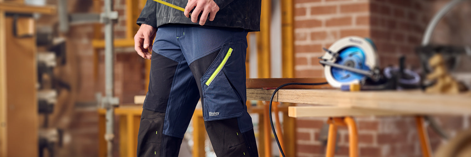 Bisley Arbeitshose in Blau mit schwarzen Einsätzen und neonfarbenem Reißverschluss, getragen in einer Werkstatt neben einer Kreissäge