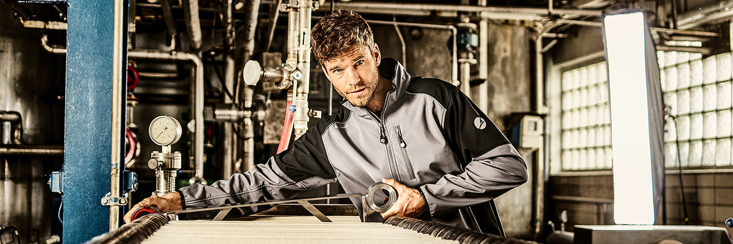 Person arbeitet an industriellen Rohrleitungen und hält ein Metallteil in der Hand, umgeben von Ventilen, Messgeräten und Maschinen in einer Werkhalle mit starkem Lichtpanel im Hintergrund