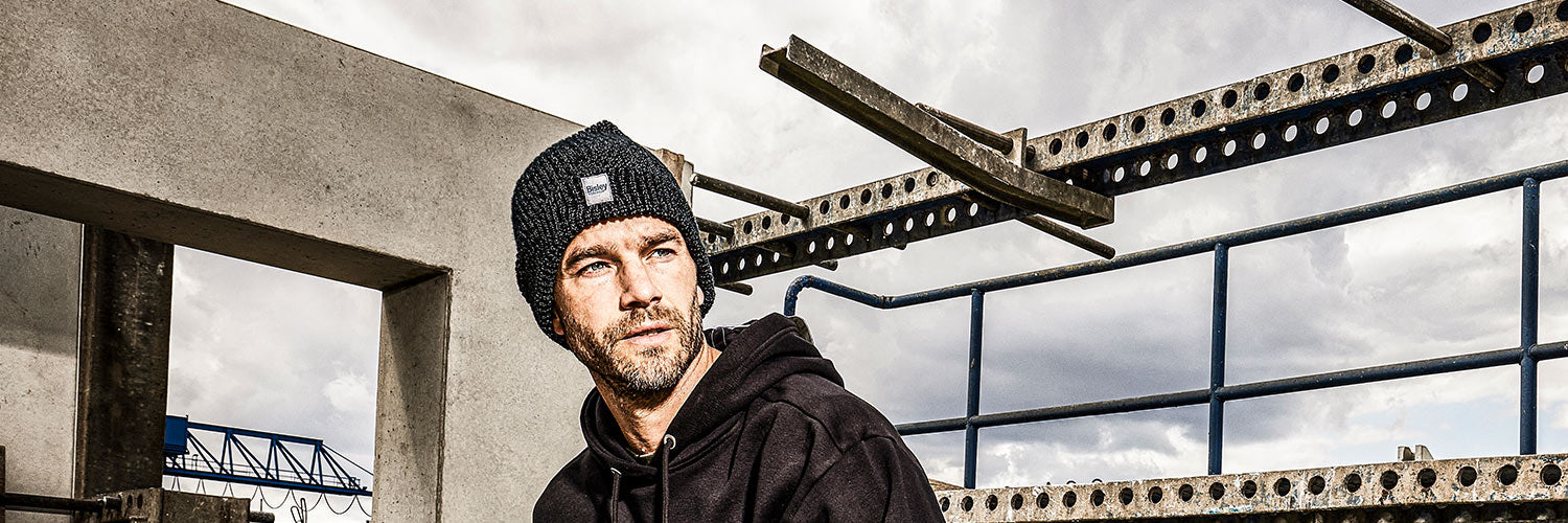 Schwarze ALBATROS Strickmütze CLAY, getragen von einer Person auf einer Baustelle mit Beton- und Stahlkonstruktionen im Hintergrund