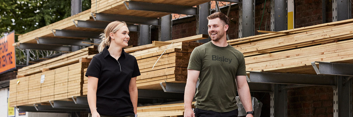 Zwei Personen auf einem Holzlagerplatz, links im schwarzen BISLEY Polo Shirt und rechts im olivgrünen BISLEY Tee T-Shirt, beide in moderner Arbeitskleidung
