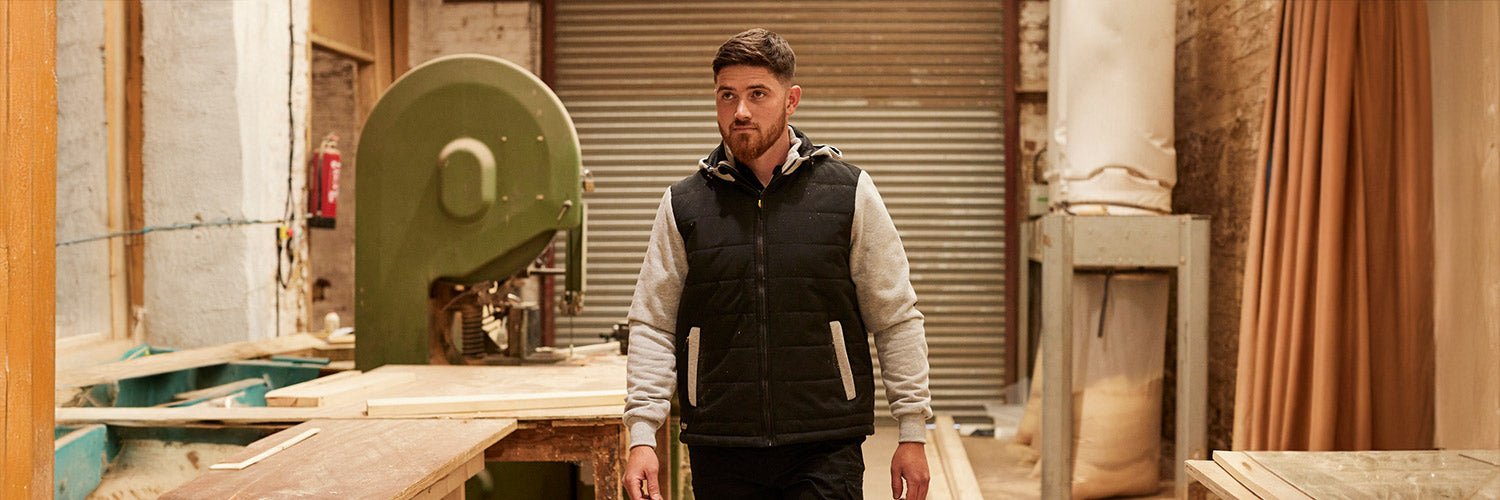 Person in Sicherheitskleidung mit schwarzer Steppweste und grauem Hoodie arbeitet in einer Werkstatt mit Holzmaschinen.