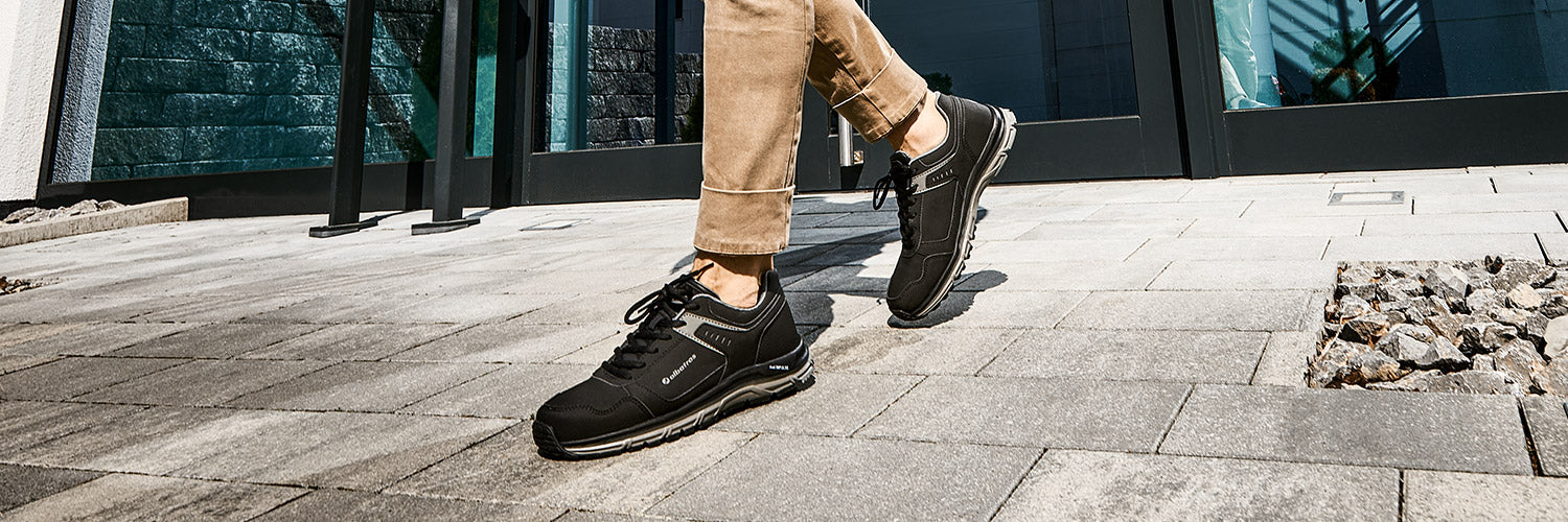 Person geht auf einem gepflasterten Außenbereich und trägt die schwarzen Berufsschuhe WHIZ ST LOW O2 ESD von ALBATROS® mit heller Sohle zu hochgekrempelter, beigefarbener Hose.