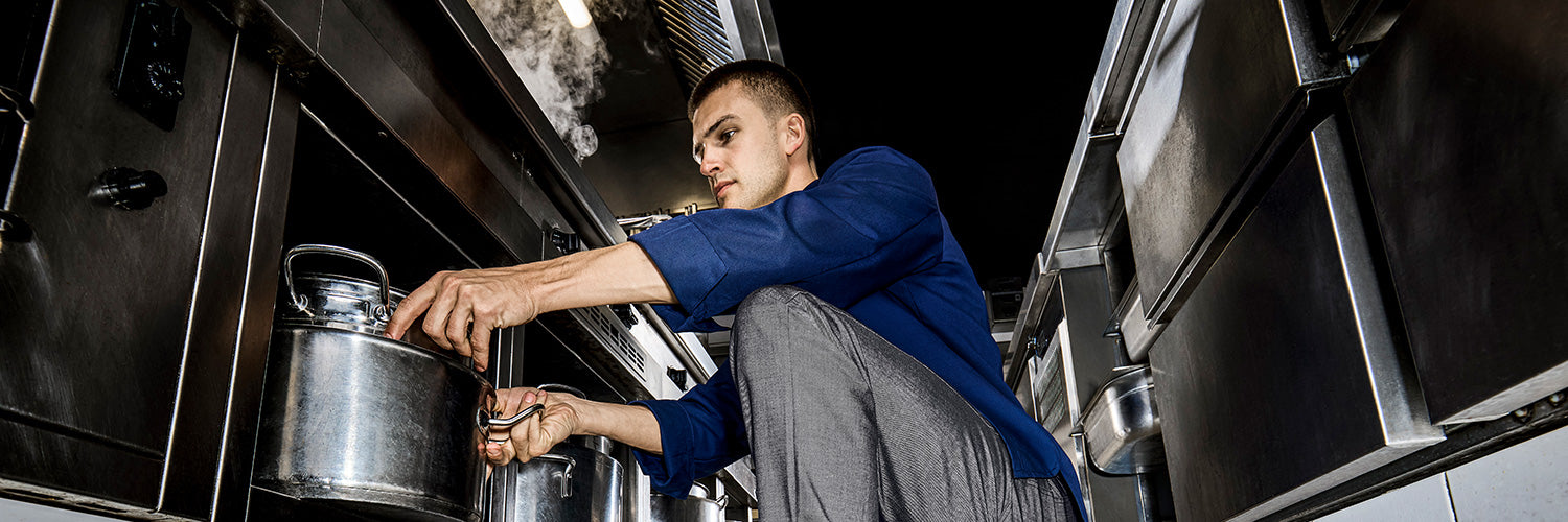 Koch in moderner Großküche trägt Arbeitskleidung mit Kochjacke und Kochhose beim Arbeiten an Edelstahlherd mit Kochtöpfen.