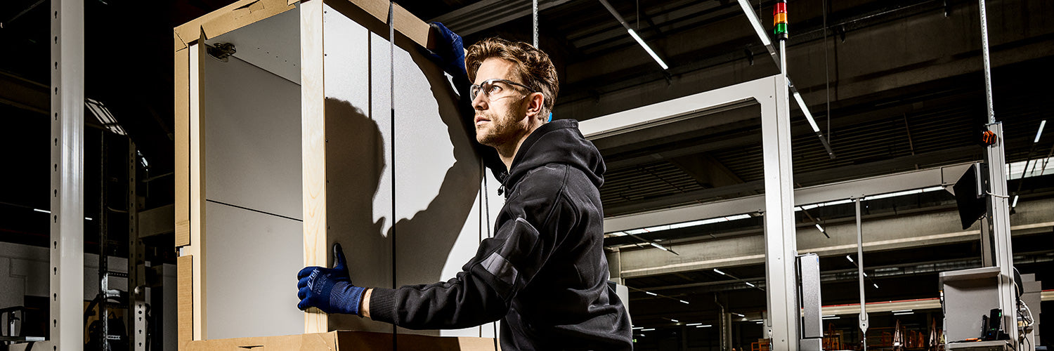 Arbeiter mit Schutzbrille, Gehörschutz und Schutzhandschuhen hebt schweres Packstück in moderner Produktionshalle mit persönlicher Schutzausrüstung.