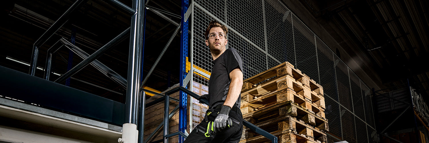 Mitarbeiter im Lager trägt persönliche Schutzausrüstung mit Schutzbrille, Arbeitshose und Schutzhandschuhen beim Arbeiten zwischen Paletten und Regalanlage.