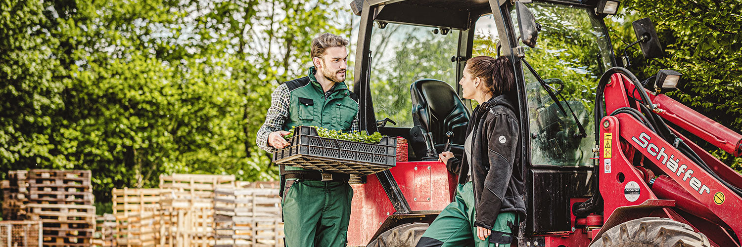 Landwirtschaftsmitarbeiter in Arbeitskleidung und persönlicher Schutzausrüstung mit Traktor und Pflanzkisten im Außenbereich.