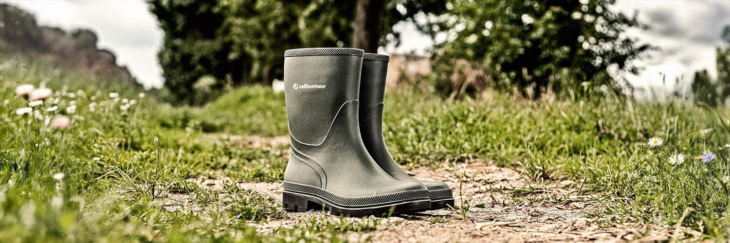 Grüne Gummistiefel von ALBATROS® steht auf einem Feldweg, umgeben von Gras und Blumen.