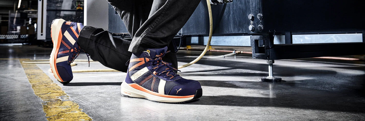 PUMA SAFETY RIVAL MID Sicherheitsschuh S1PS ESD in Blau mit orangefarbenen Details, getragen von einer Person auf Industrieboden.