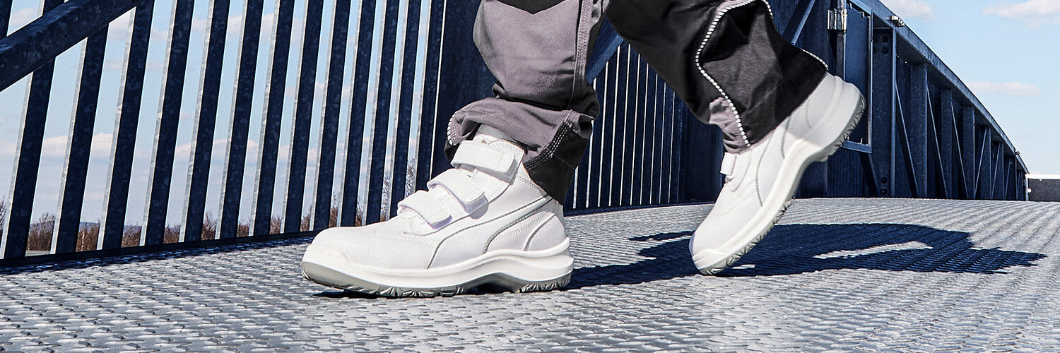 PUMA SAFETY ABSOLUTE MID Sicherheitsschuh S2 in Weiß mit Klettverschluss, getragen von einer Person auf Metallsteg im Freien.