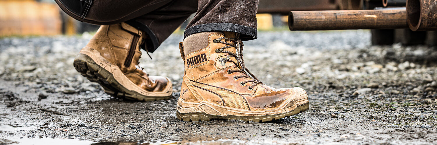 PUMA SAFETY CONQUEST HIGH Sicherheitsschuh S3 in Braun, getragen von einer Person im Outdoor-Einsatz auf matschigem Boden.