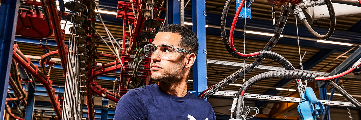 Arbeiter in industrieller Umgebung trägt eine transparente Schutzbrille zum Schutz der Augen. Im Hintergrund sind Maschinen, Leitungen und Metallstrukturen zu sehen.