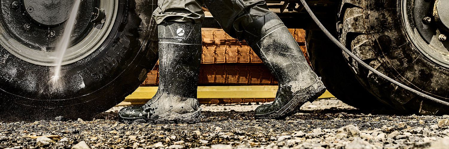 Schwarzer Sicherheitsgummistiefel von ALBATROS, getragen von einer Person beim Arbeiten auf nassem Schotterboden neben großem Reifen mit Wasserstrahl.