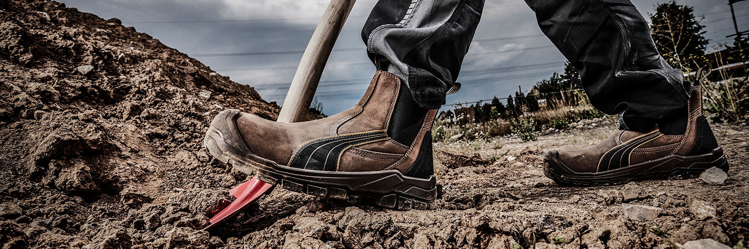 Arbeiter steht auf einer Baustelle in unebenem Erdreich und trägt die braunen Sicherheitsschuhe TANAMI MID S3 von PUMA SAFETY® im robusten Stiefel-Design.