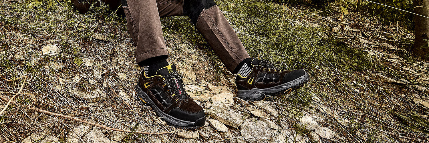 Person sitzt im Gelände auf steinigem Boden mit Grasbewuchs und trägt den Trekkingstiefel LOFOTEN 2.0 BROWN CTX LOW von ALBATROS® zu brauner Outdoorhose.