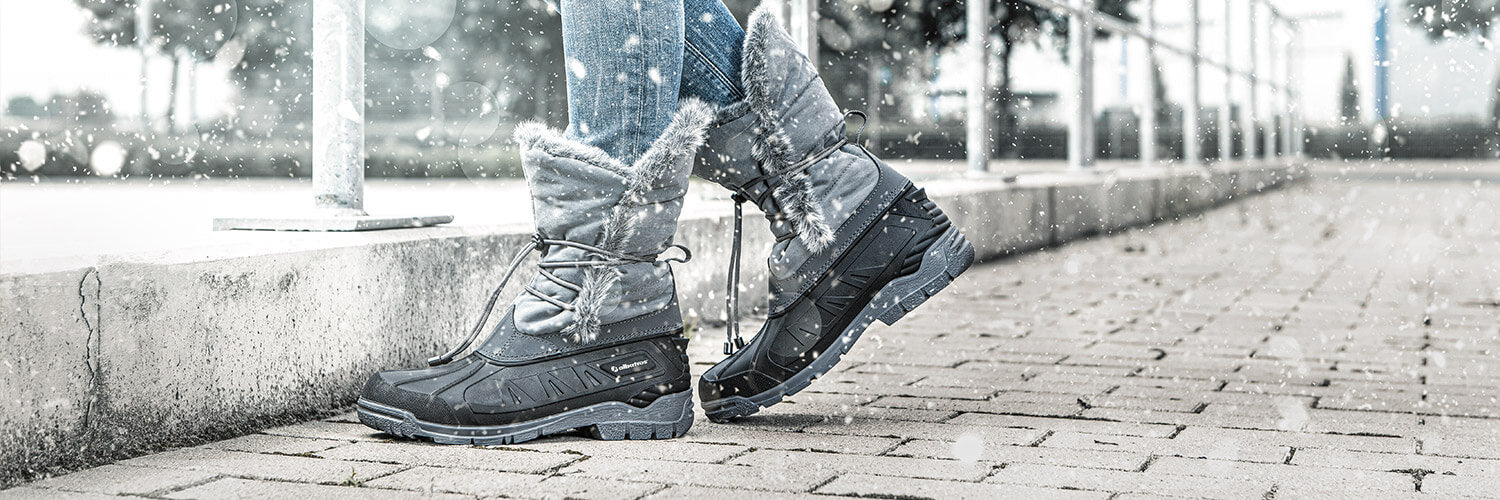 Person trägt schwarze Winterstiefel mit grauem, pelzgefüttertem Schaft und Schnürung, geht auf gepflastertem Weg im Schneefall.