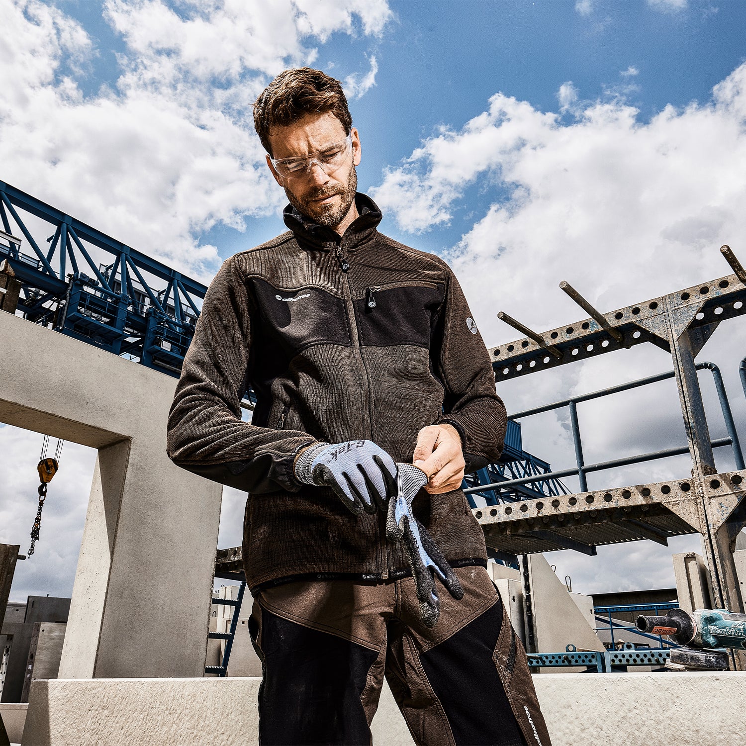 Mann in brauner Arbeitskleidung von Albatros und transparenter Schutzbrille zieht Arbeitshandschuhe von G-Tek auf einer Baustelle an.