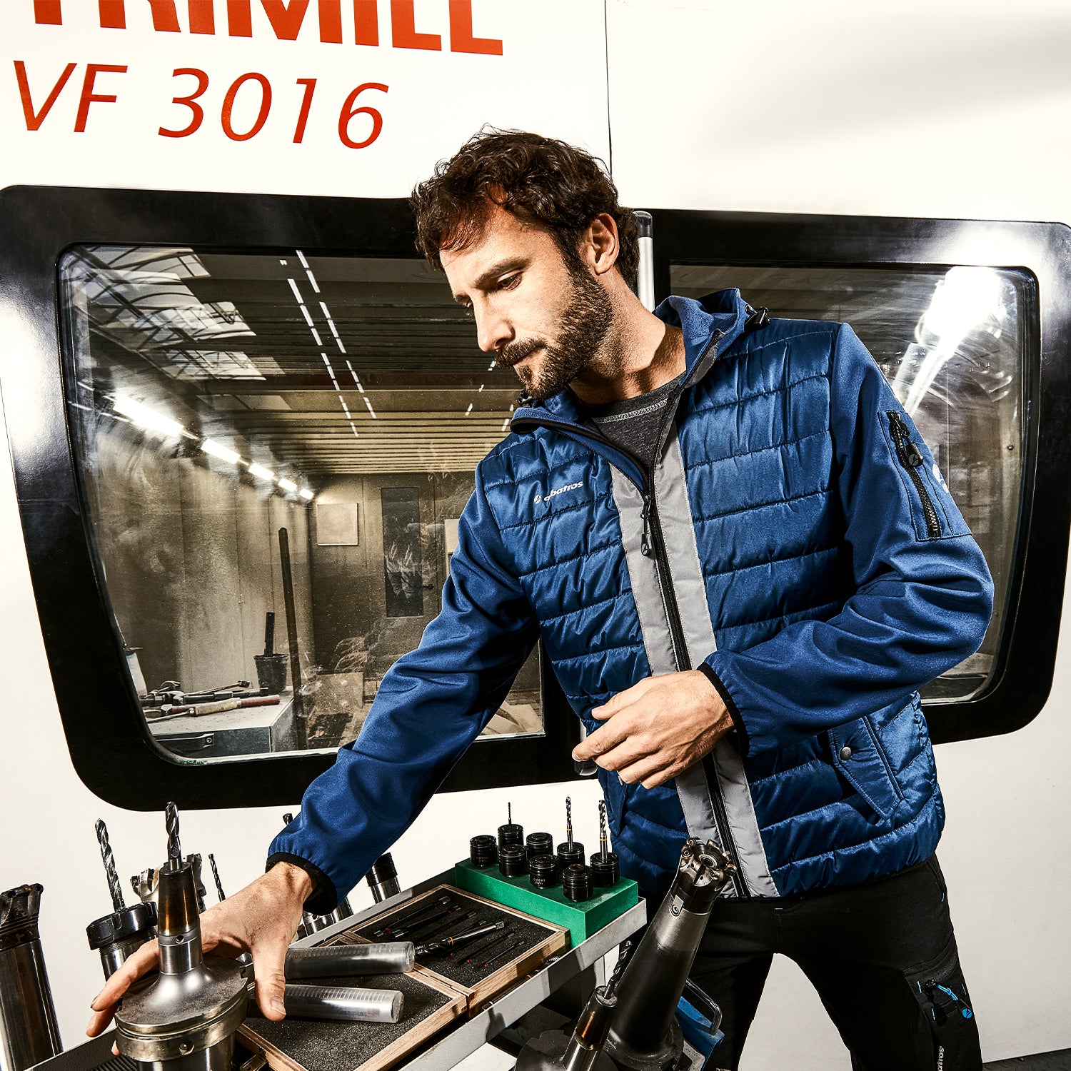 Mann in blauer Arbeitsjacke von Albatros bedient Werkzeuge vor einer CNC-Maschine.